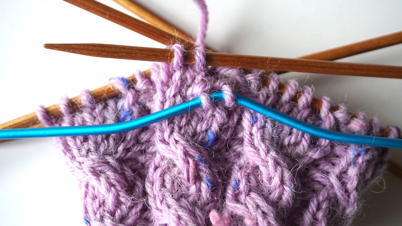 Step-by-step knitting photo showing purple knitting project with two stitches on a blue cable needle