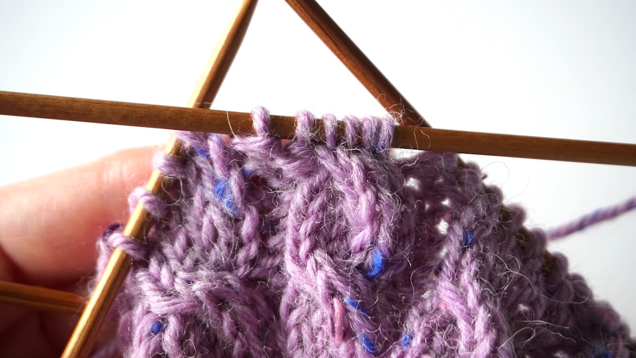 Step-by-step knitting photo showing purple knitting project with finished cable knit stitch pattern