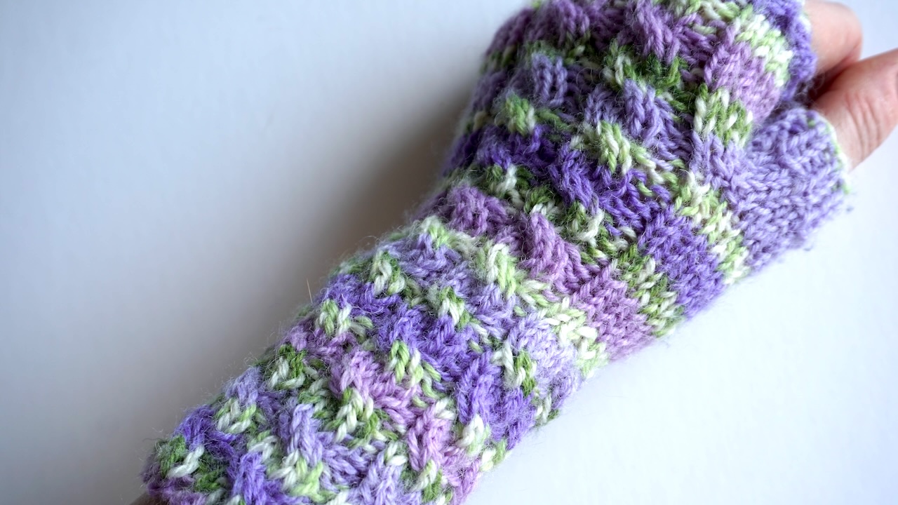 Hand wearing a fingerless mitten featuring cable knit pattern in purple and green hued yarn