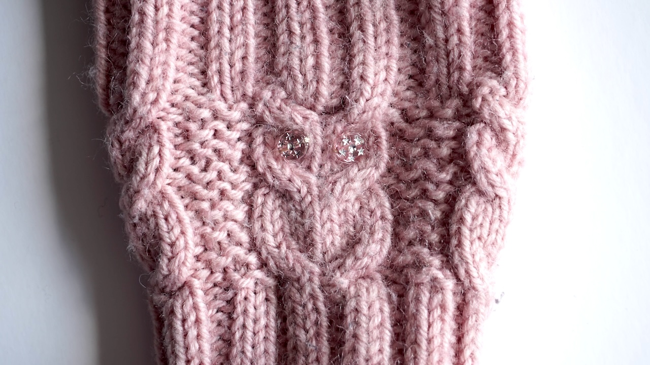 Close-up photo of cable knit stitch texture in light pink yarn showing buttons as owl's eyes