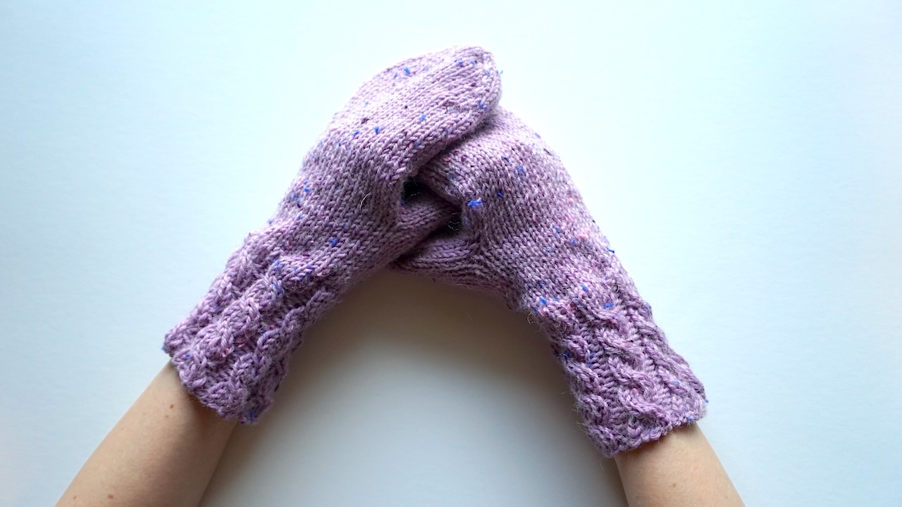 Hands wearing knitted mittens featuring cable cuffs in light purple yarn