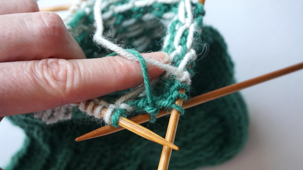 Close-up knitting photo showing fingers twisting different colored yarns on the wrong side of a colorwork pattern