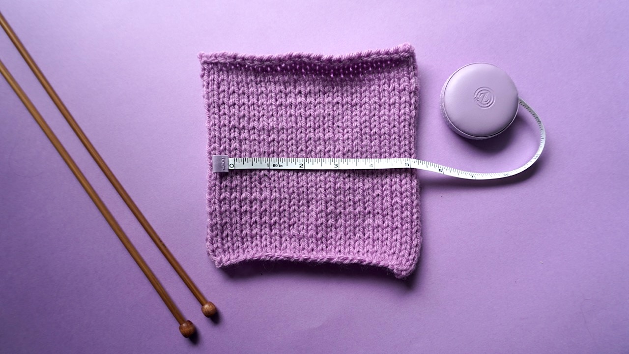 A purple knitting gauge swatch with a tape measure on top of it and wooden knitting needles laying on the table