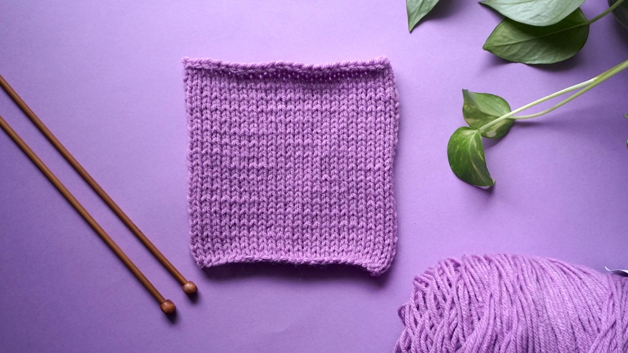 A purple knitting gauge swatch with wooden knitting needles, yarn and a house plant laying next to it