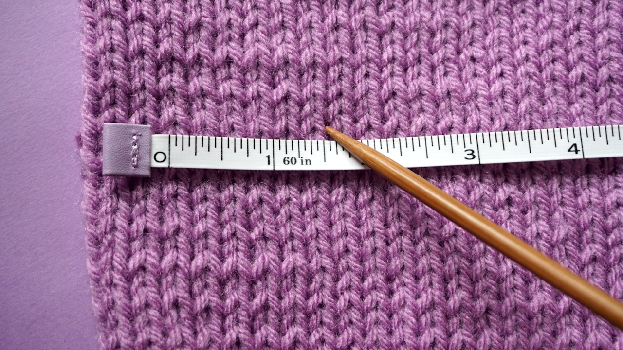 A tape measure on top of a purple knitting gauge swatch with a wooden needle pointing at a knit stitch