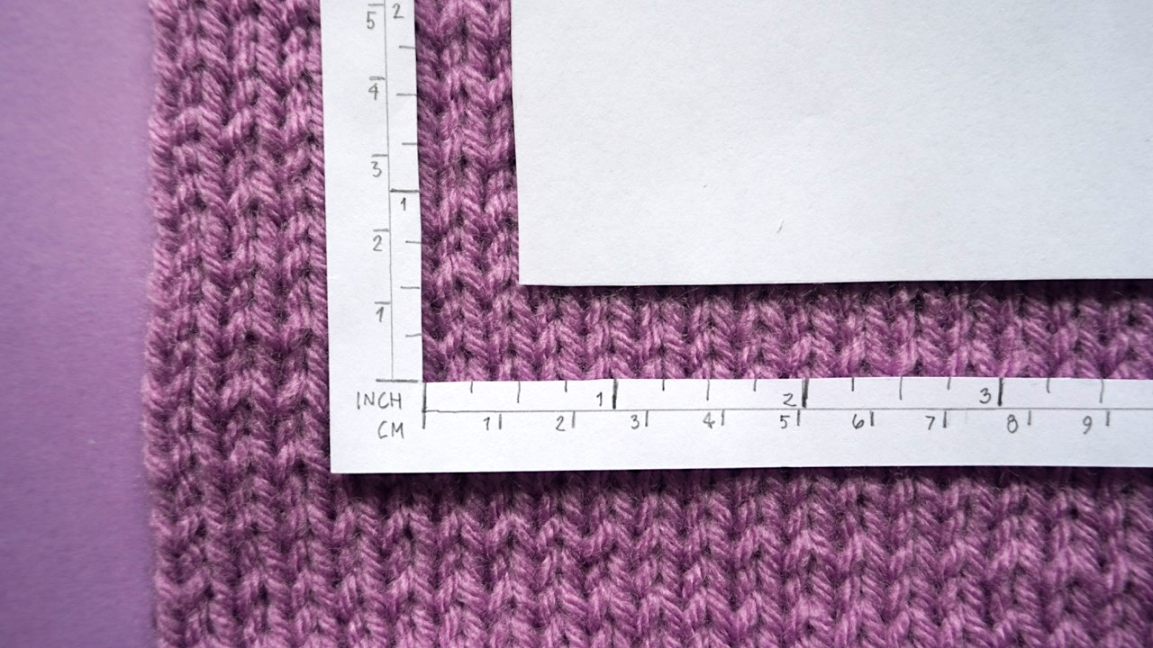A knitting gauge tool laying on top of a purple gauge swatch