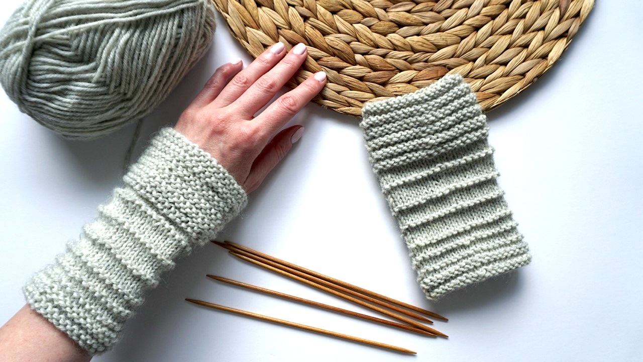 Hand wearing a knitted hand warmer in light gray yarn next to another hand warmer, knitting needles, and a skein of yarn