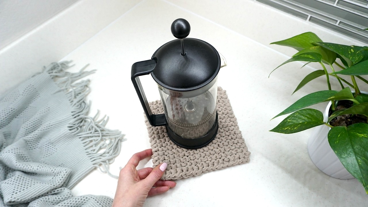 A French press on top of a beige knitted trivet next to a throw and a potted plant