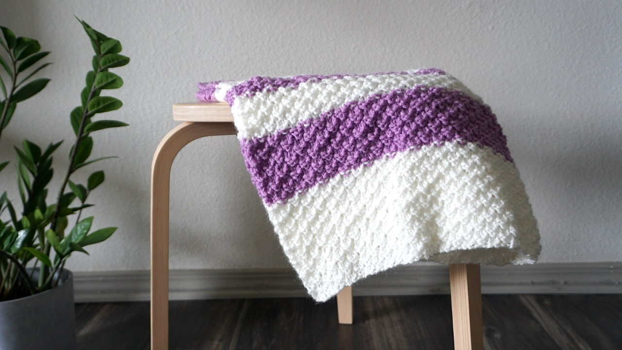 A knitted baby blanket in double moss stitch and white and purple color blocks laying on top of a stool