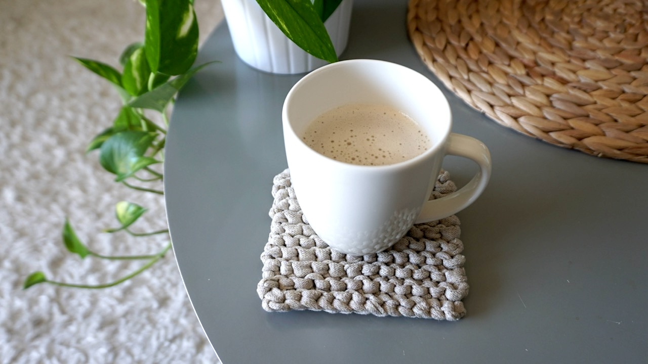 A white coffee cup on top of a beige knitted trivet next to a potted plant