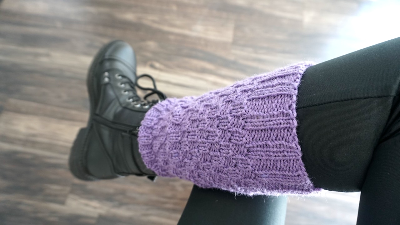 A foot wearing a purple knitted leg warmer in ribbed checks pattern and black boot