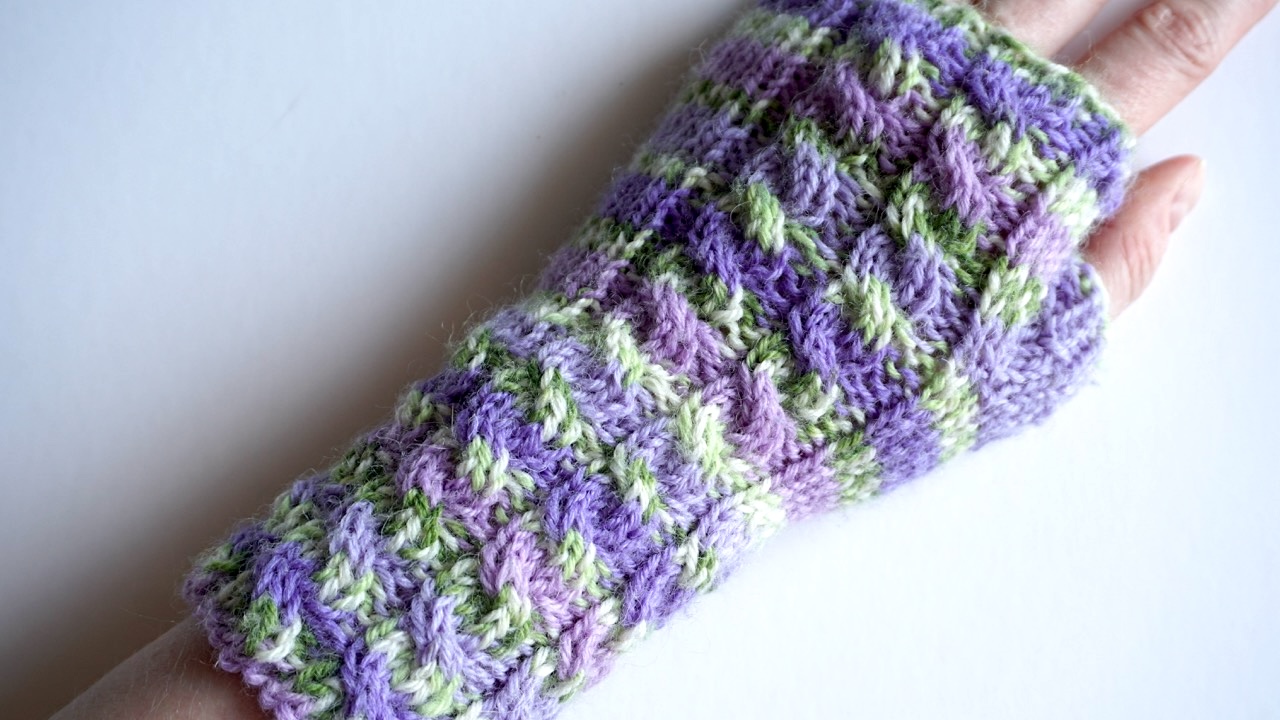 A hand wearing a knit fingerless glove featuring cables in soft purple and green yarn