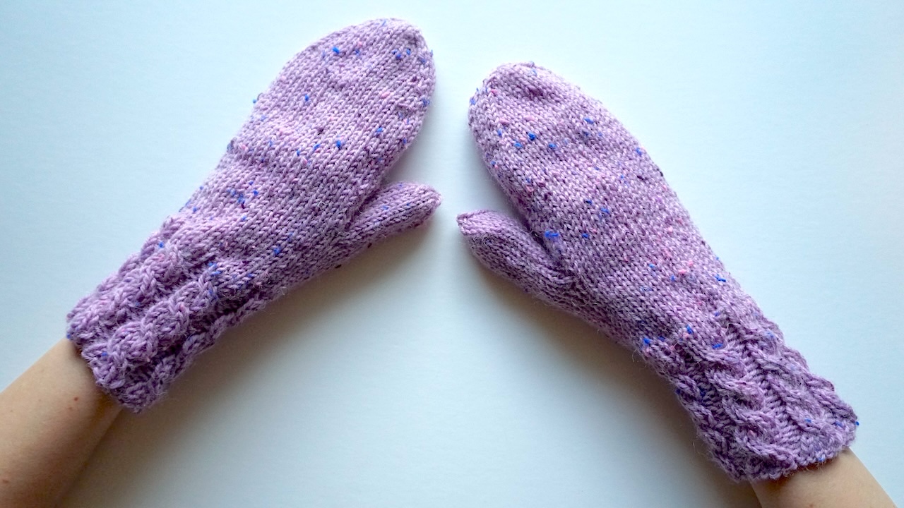 Hands wearing knitted mittens featuring cable cuffs in light purple yarn