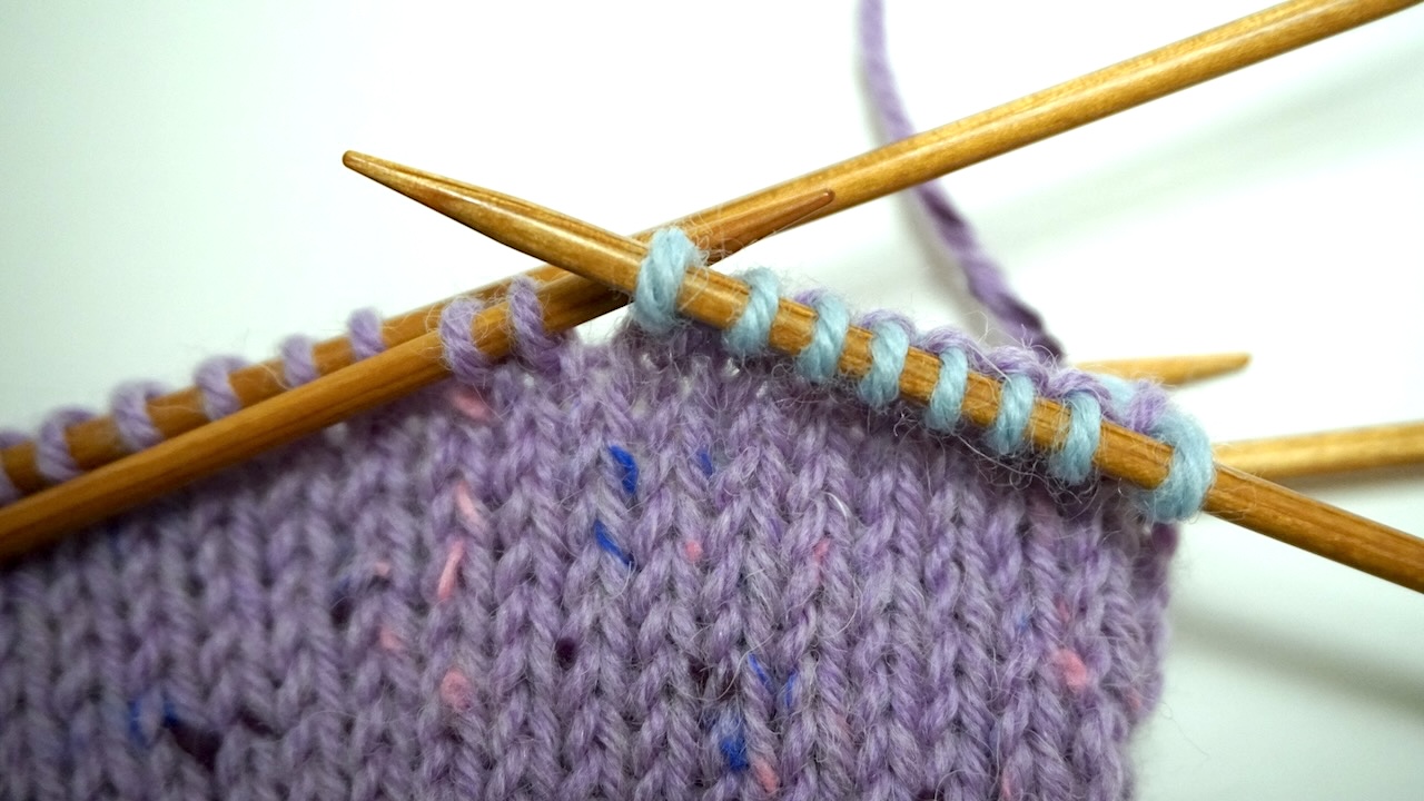 Step-by-step knitting photo showing light blue thumb stitches on the right needle and purple stitches on the left needle
