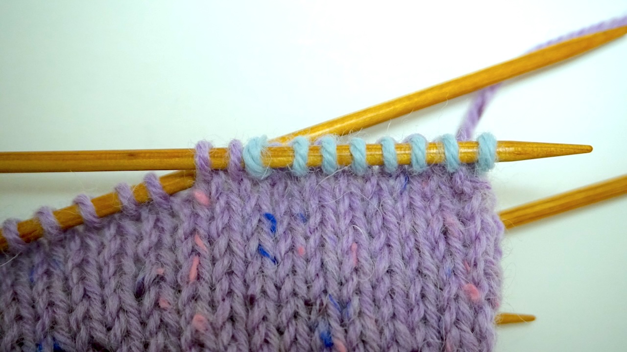 Step-by-step knitting photo showing light blue thumb stitches next to purple stitches on the left needle