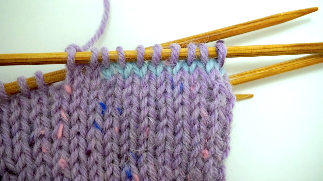 Step-by-step knitting photo showing light blue thumb stitches next knitted with purple working yarn