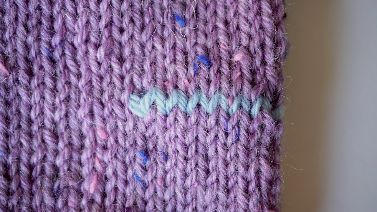 Close-up photo of the thumb hole stitches kitted in light blue yarn on a purple mitten