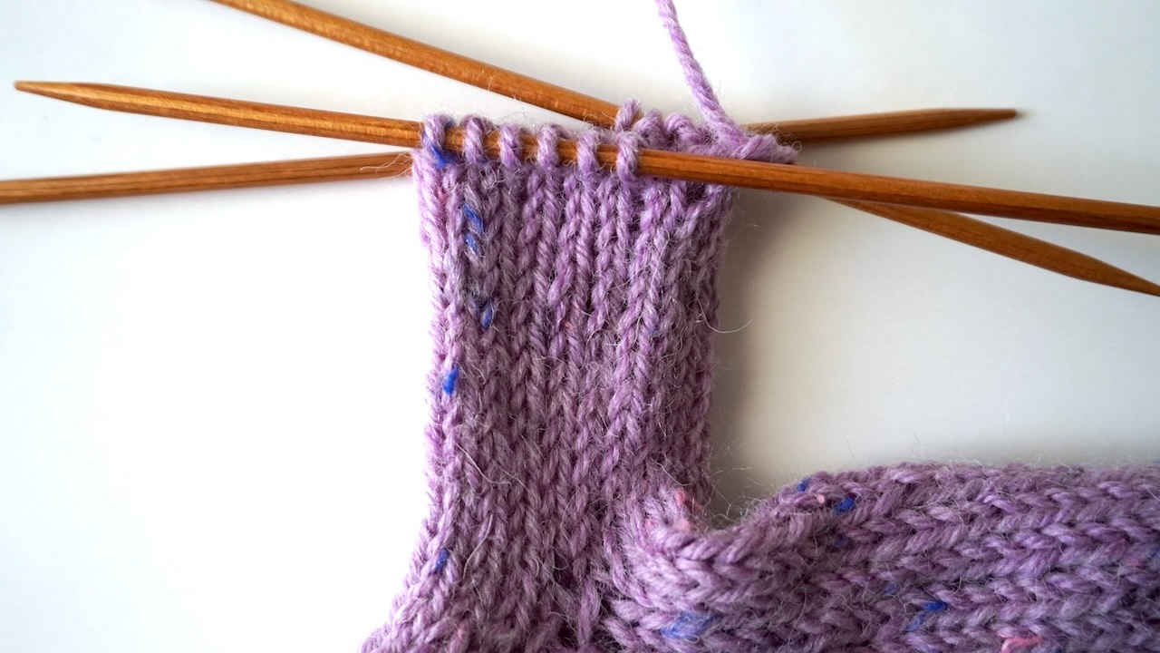 Step-by-step knitting photo showing the body of the thumb featuring stockinette stitch in purple yarn
