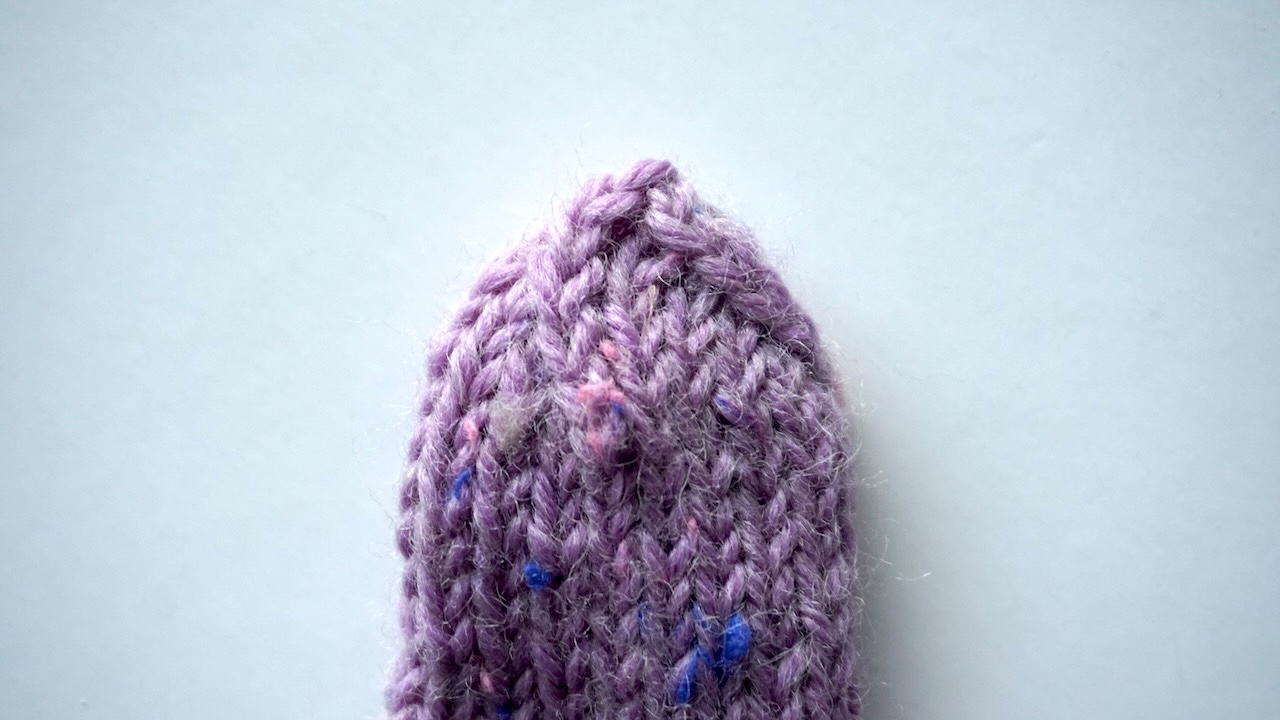 Step-by-step knitting photo showing the finished mitten thumb featuring stockinette stitch in purple yarn