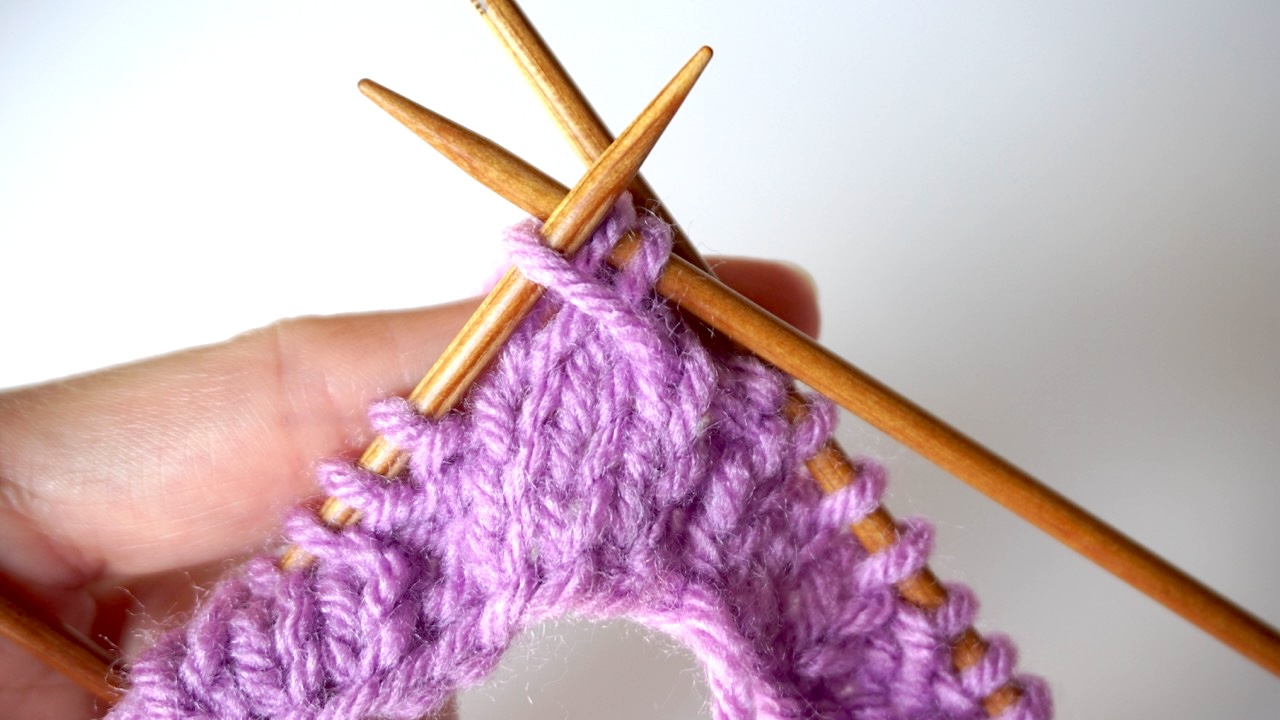 Step-by-step knitting photo showing hands lifting a slipped stitch over two knit stitches for mock cable stitch