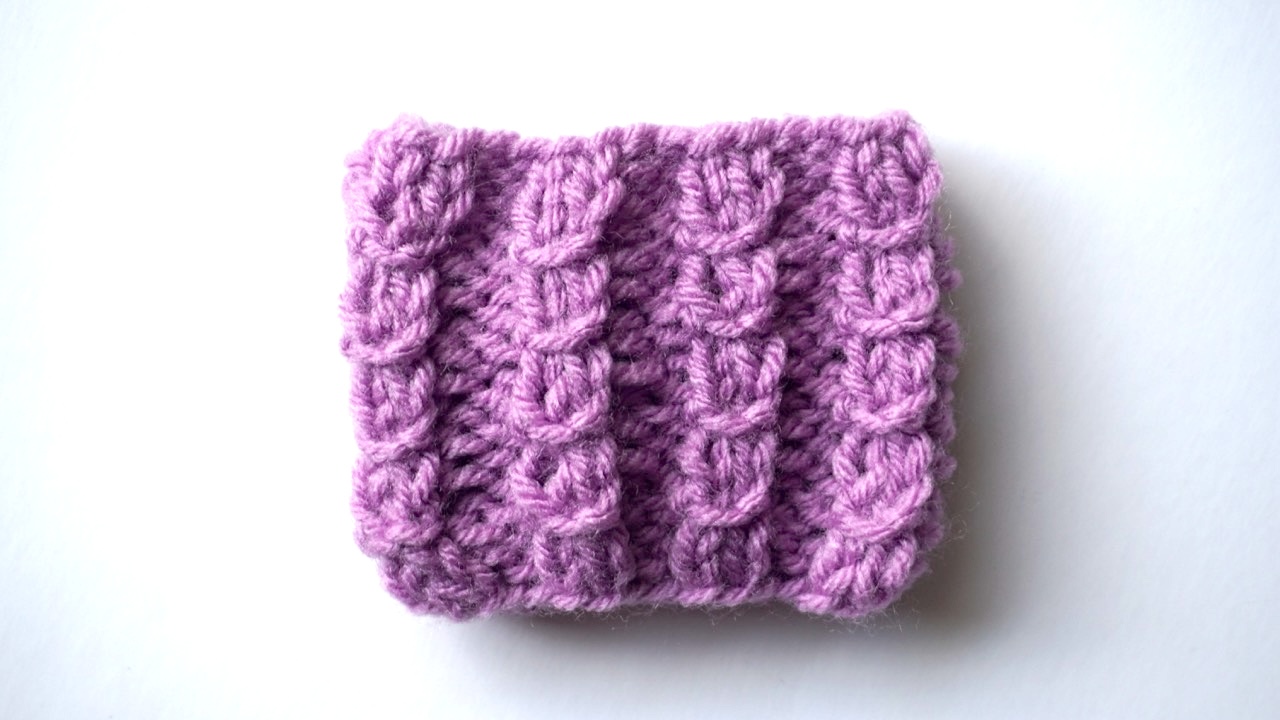 A purple knitting swatch showing mock cable with a yarn loop pattern