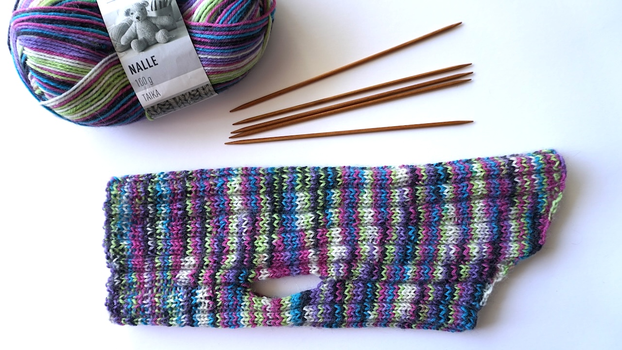 Knit dog sweater in multicolored stripes next to double pointed needles and s skein of yarn