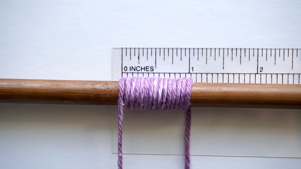 Purple yarn wrapped around a knitting needle and a tape measure measuring the wraps per inch in order to find out the yarn weight